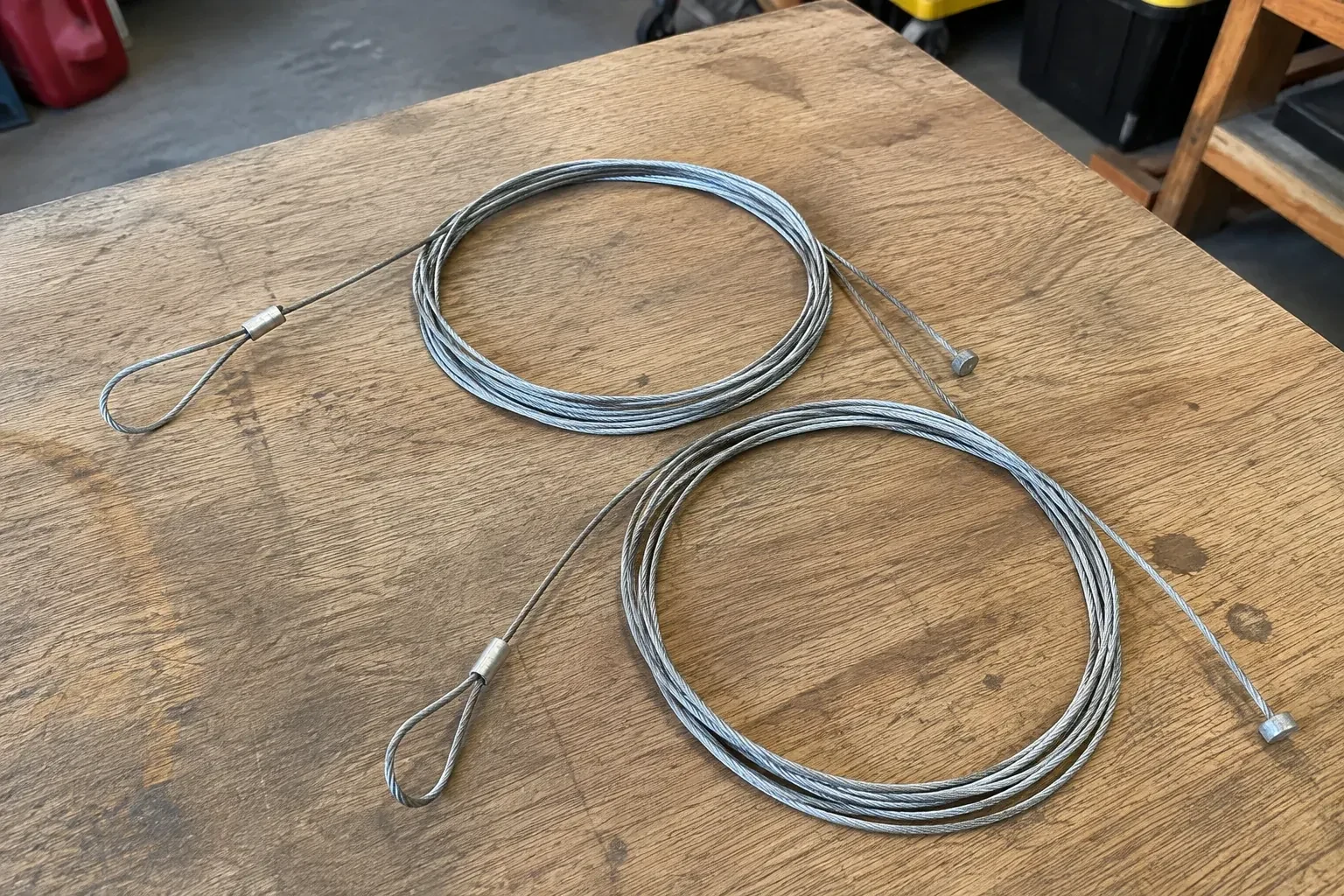 Two coiled steel cables with loop and stop ends on a workbench. — Garage Door Cable Replacement, Islington